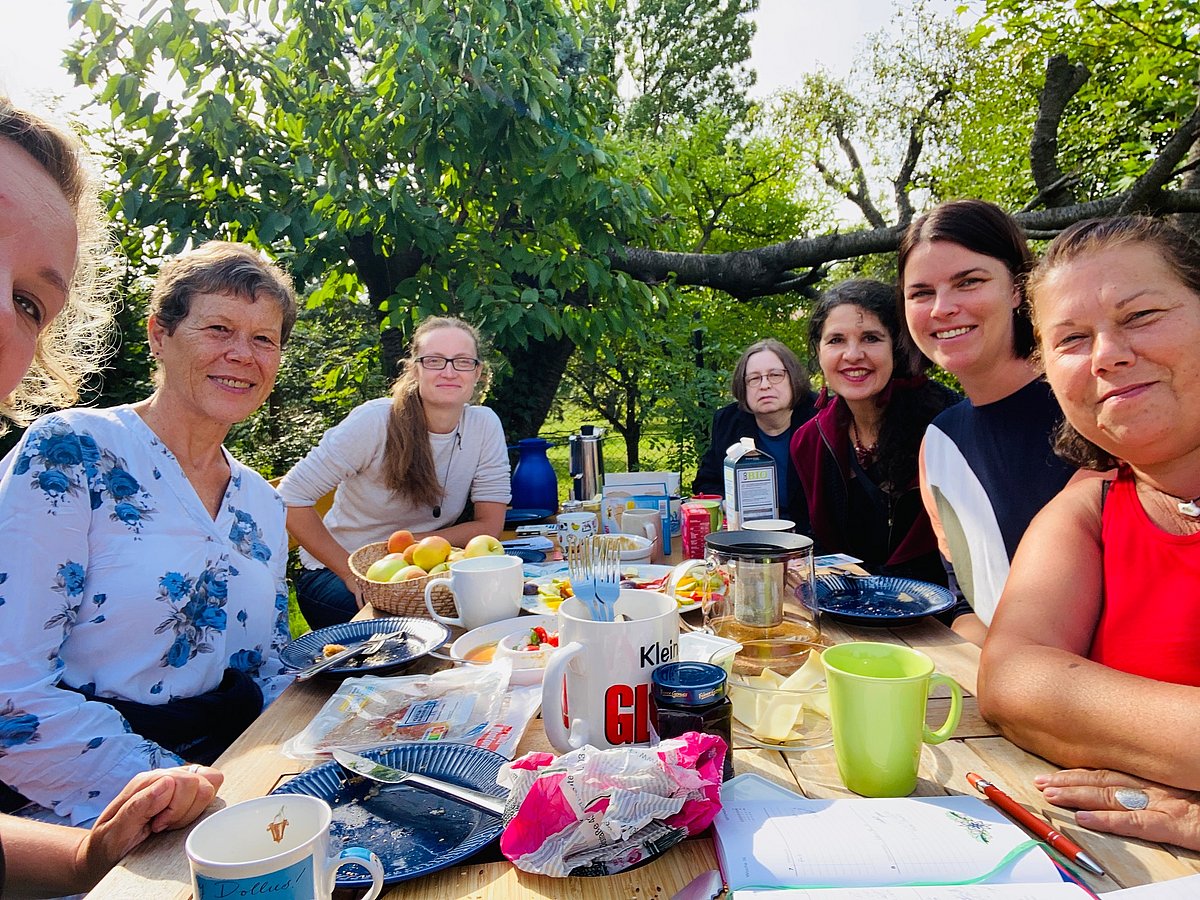 Sieben Frauen sitzen an einem Frühstücktisch im Garten und schauen in die Kamera