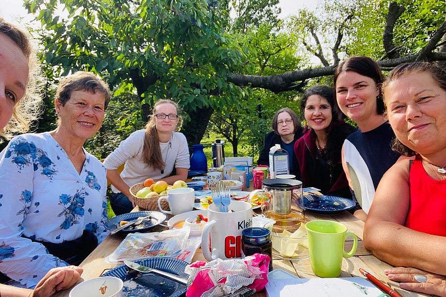 Sieben Frauen sitzen an einem Frühstücktisch im Garten und schauen in die Kamera
